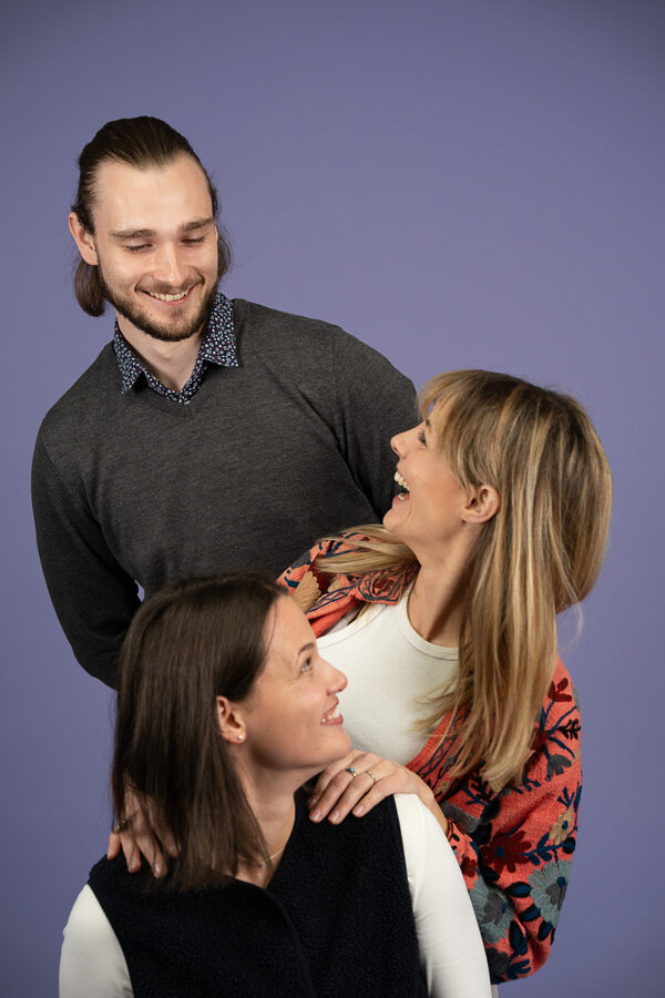 Leo, Vikky und Jana von Mind The Brain vor violettem Hintergrund, eng beieinander und gemeinsam lachend